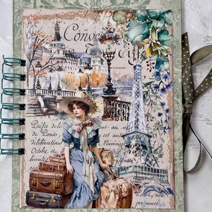 Può includere: Un diario a spirale con un tema parigino vintage. La copertina presenta un'illustrazione ad acquerello di una donna con bagagli, la Torre Eiffel ed elementi architettonici. Il testo include la parola "Conve" e altri testi francesi.