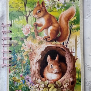 Könnte beinhalten: Ein Spiralnotizbuch mit einer bezaubernden Illustration von zwei Eichhörnchen in einer Waldszene. Das Cover zeigt ein braunes Eichhörnchen auf einem Ast und ein weiteres in einer Baumhöhle, umgeben von Blumen und üppigem Grün.