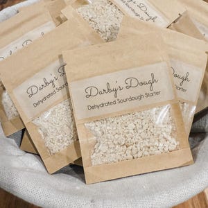 Puede incluir: Primer plano de varias bolsas peque&ntilde;as de papel marr&oacute;n llenas de masa madre blanca. Cada bolsa tiene una etiqueta que dice "Darby's Dough Masa madre de masa madre deshidratada".