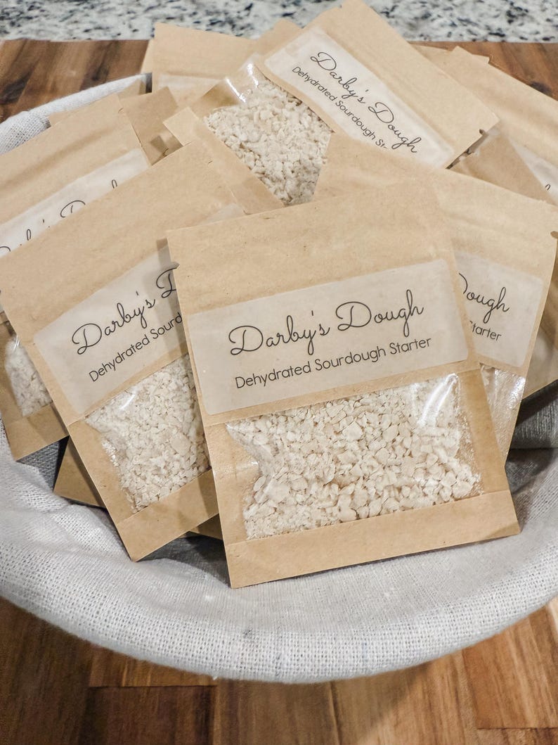 Puede incluir: Primer plano de varias bolsas peque&ntilde;as de papel marr&oacute;n llenas de masa madre blanca. Cada bolsa tiene una etiqueta que dice "Darby's Dough Masa madre de masa madre deshidratada".
