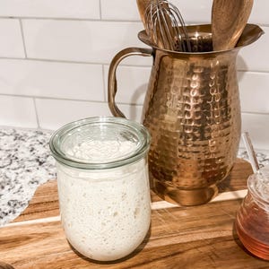 Puede incluir: Un tarro de vidrio con un l&iacute;quido blanco y burbujeante en su interior, sentado sobre una tabla de cortar de madera. Detr&aacute;s del tarro hay una jarra de cobre con cucharas de madera y un batidor dentro. Un peque&ntilde;o tarro de vidrio con un l&iacute;quido de color miel est&aacute; a la derecha del tarro.