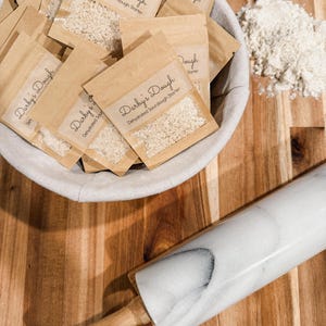Puede incluir: Un bol blanco lleno de peque&ntilde;as bolsas de papel marr&oacute;n que contienen masa madre deshidratada. Las bolsas est&aacute;n etiquetadas con "Darby's Dough Masa madre de masa madre deshidratada". Un rodillo de m&aacute;rmol blanco y un bol de harina est&aacute;n sobre una superficie de madera.