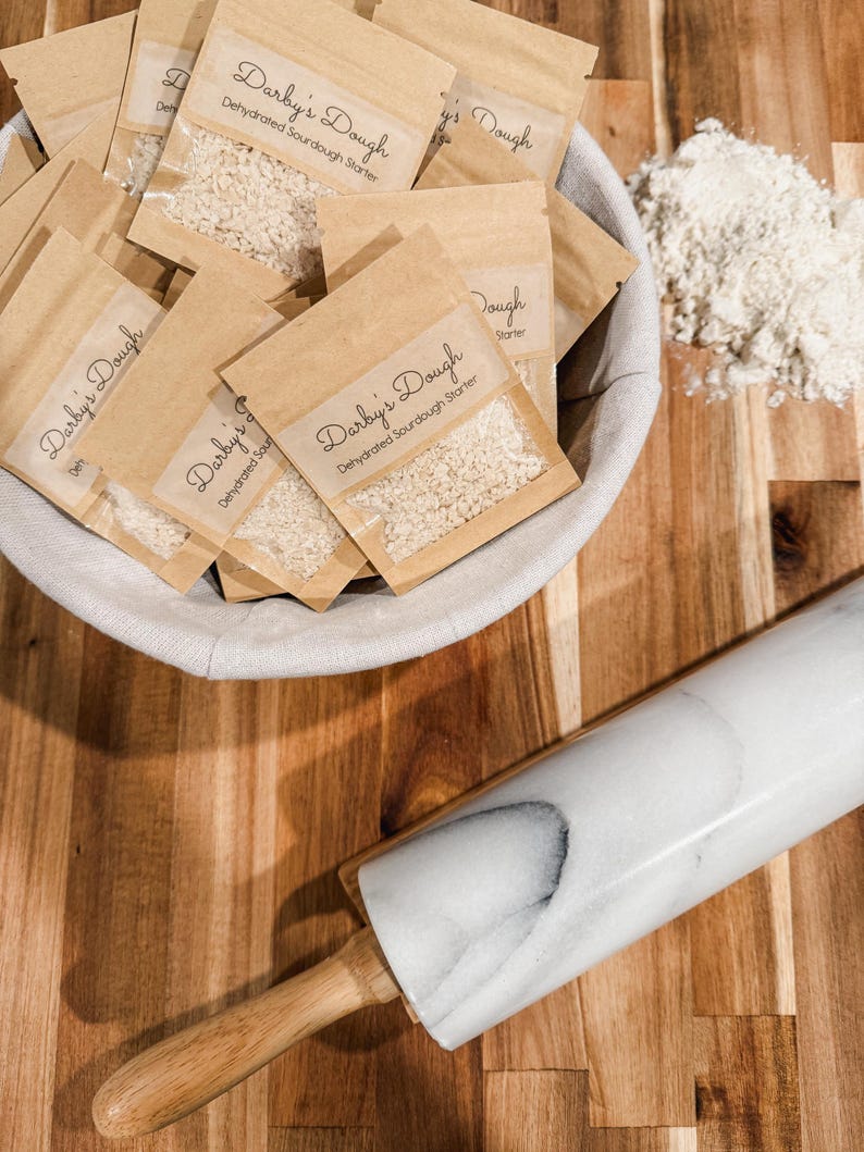 Puede incluir: Un bol blanco lleno de peque&ntilde;as bolsas de papel marr&oacute;n que contienen masa madre deshidratada. Las bolsas est&aacute;n etiquetadas con "Darby's Dough Masa madre de masa madre deshidratada". Un rodillo de m&aacute;rmol blanco y un bol de harina est&aacute;n sobre una superficie de madera.