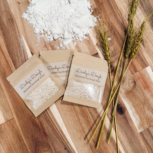 Puede incluir: Tres peque&ntilde;os paquetes de papel marr&oacute;n con el texto "Darby's Dough Dehydrated Sourdough Starter" en ellos. Los paquetes contienen un polvo blanco. En el fondo hay un mont&oacute;n de harina blanca, junto con tres tallos de trigo.