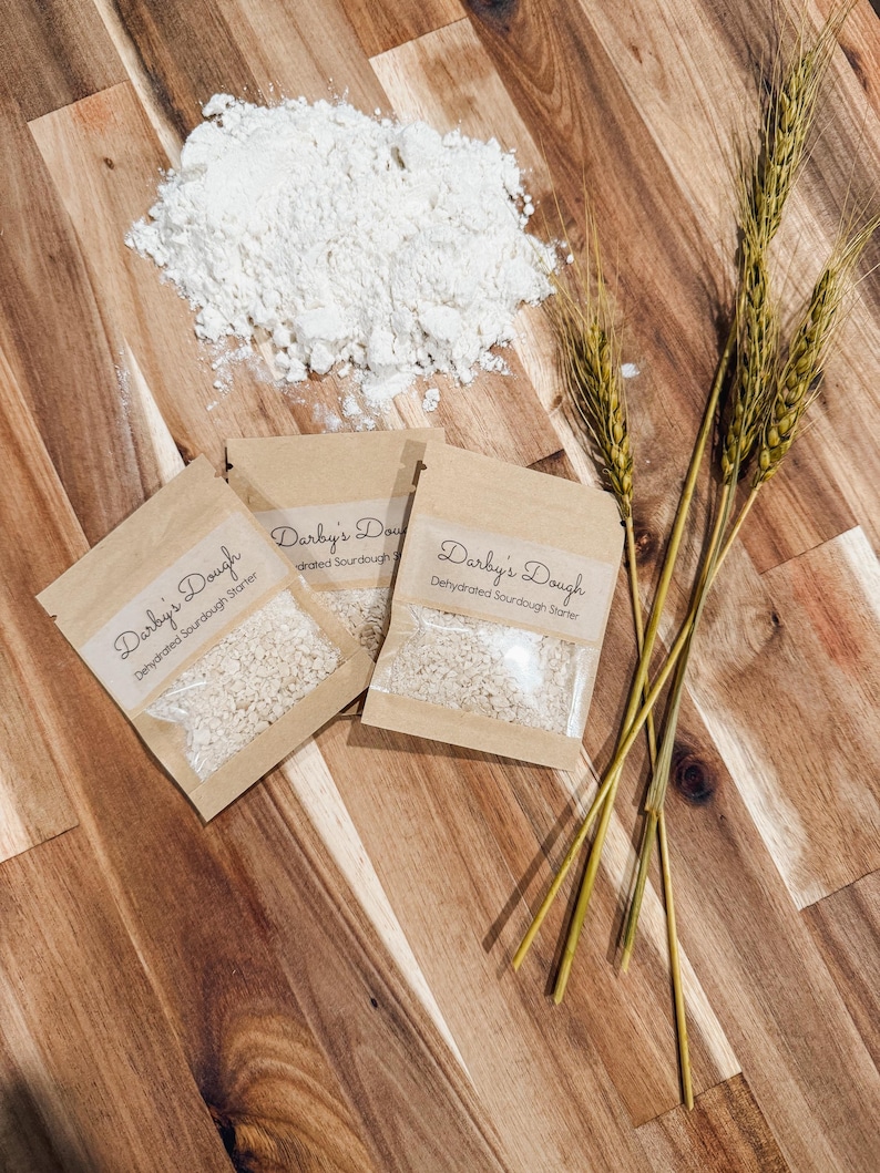 Puede incluir: Tres peque&ntilde;os paquetes de papel marr&oacute;n con el texto "Darby's Dough Dehydrated Sourdough Starter" en ellos. Los paquetes contienen un polvo blanco. En el fondo hay un mont&oacute;n de harina blanca, junto con tres tallos de trigo.