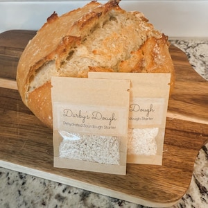 May include: Two small brown paper packets containing dehydrated sourdough starter. The packets are labelled "Darby's Dough Dehydrated Sourdough Starter" and "Dough dough starter". A loaf of freshly baked sourdough bread is in the background.