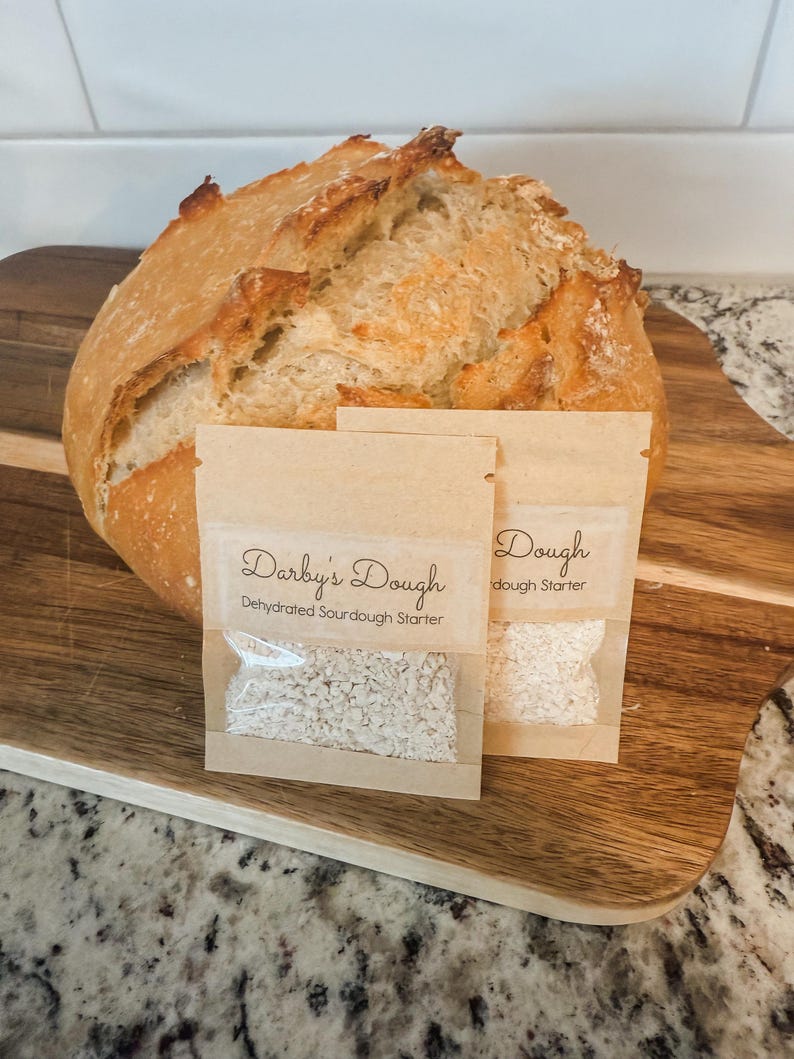 Puede incluir: Dos peque&ntilde;os paquetes de papel marr&oacute;n que contienen masa madre deshidratada. Los paquetes est&aacute;n etiquetados como "Darby's Dough Masa madre deshidratada" y "Dough masa madre". Un pan de masa madre reci&eacute;n horneado est&aacute; en el fondo.