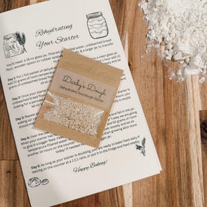 Puede incluir: Instrucciones para rehidratar un fermento de masa madre con un paquete de fermento de masa madre deshidratado. Las instrucciones incluyen los pasos para alimentar el fermento y una nota sobre la felicidad de hornear.