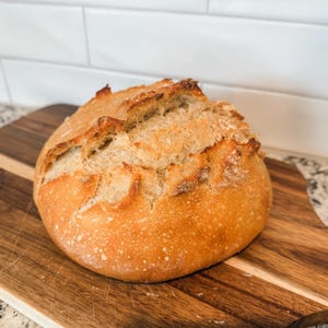 Puede incluir: Una hogaza de pan de masa madre reci&eacute;n horneada con una corteza dorada y una parte superior ligeramente agrietada. El pan est&aacute; sobre una tabla de cortar de madera.