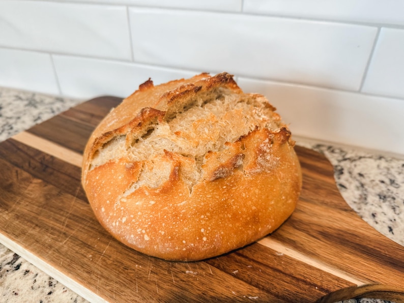Puede incluir: Una hogaza de pan de masa madre reci&eacute;n horneada con una corteza dorada y una parte superior ligeramente agrietada. El pan est&aacute; sobre una tabla de cortar de madera.