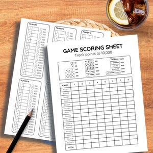 Puede incluir: Un conjunto de hojas blancas para anotar puntuaciones de juegos, con texto negro y un lápiz. Las hojas están diseñadas para rastrear puntos hasta 10.000, con espacios para varios jugadores y rondas. Un vaso de té helado con una rodaja de limón en el fondo.