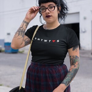 May include: A black t-shirt with the text "SMTWT&hearts;S" printed in white. The person wearing the shirt has a tattoo on their arm.