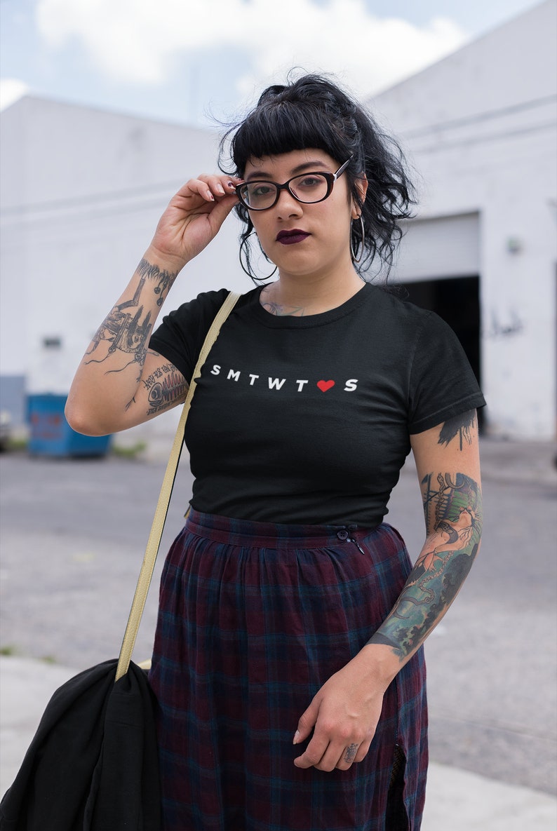 May include: A black t-shirt with the text "SMTWT&hearts;S" printed in white. The person wearing the shirt has a tattoo on their arm.