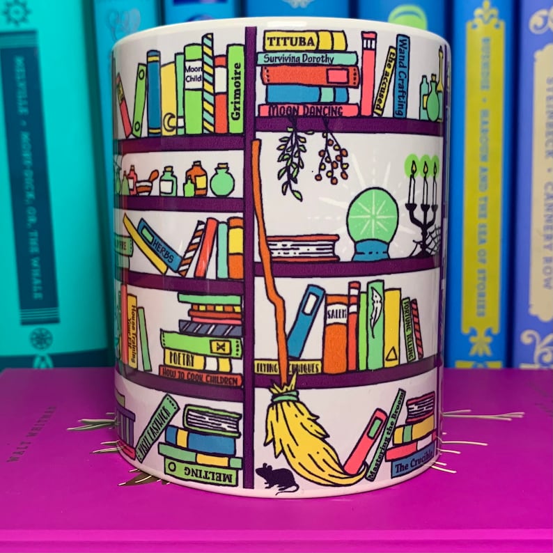May include: A white ceramic mug with a colorful illustration of a bookshelf filled with books, a broom, a crystal ball, and a mouse. The books have titles like "Grimoire", "Moon Dancing", "Tituba", and "Wand Crafting".