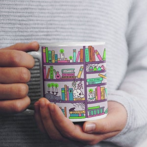 May include: White ceramic mug with a colorful illustration of a bookshelf filled with books, potions, and other magical items. The books have titles like "Diary", "Spells", "Hex in the City", "Toil and Trouble", "Love Potions", "Trial by Fire", "Hamlet", and "How to Turn Men into Toads!"