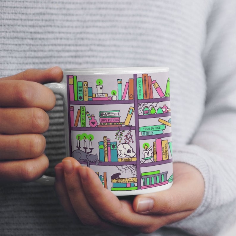 May include: White ceramic mug with a colorful illustration of a bookshelf filled with books, potions, and other magical items. The books have titles like "Diary", "Spells", "Hex in the City", "Toil and Trouble", "Love Potions", "Trial by Fire", "Hamlet", and "How to Turn Men into Toads!"