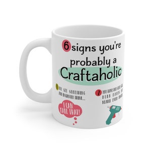 May include: White ceramic mug with a red and green design. The mug features the text "6 signs you're probably a Craftaholic" and lists signs like "You see something and instantly think... I can make that!" and "You have hot glue burn marks on nearly every finger".