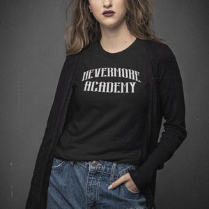 May include: A woman wearing a black long-sleeved cardigan over a black t-shirt with the text "NEVERMORE ACADEMY" in white. She is wearing blue jeans and has her hand in her pocket.