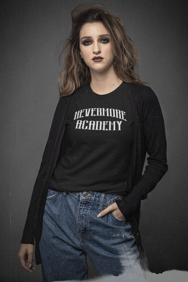 May include: A woman wearing a black long-sleeved cardigan over a black t-shirt with the text "NEVERMORE ACADEMY" in white. She is wearing blue jeans and has her hand in her pocket.