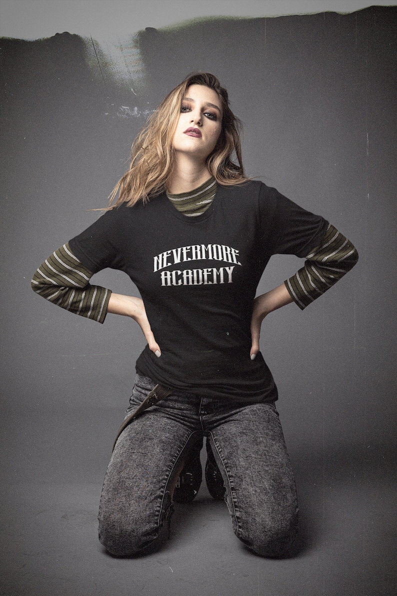 May include: A woman wearing a black long-sleeved shirt with the text "NEVERMORE ACADEMY" printed on the front. She is also wearing a green and black striped shirt underneath and dark gray jeans.