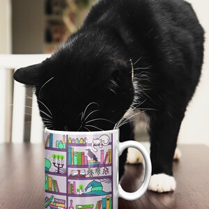 May include: A white ceramic mug with a black cat peeking into it. The mug features a colorful illustration of a bookshelf filled with books, potions, and other magical items. The text "Wicked" is visible on one of the books.