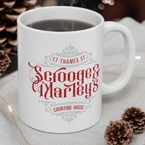 May include: White ceramic mug with a red and gray design. The mug features the text "Scrooge & Marley's" with the address "17 Thames St." and "Counting House" underneath.