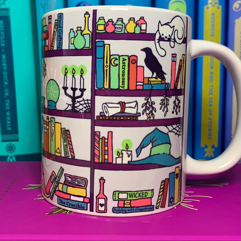 May include: White ceramic mug with a colorful illustration of a bookshelf filled with books, potions, candles, and other magical items. The text on the books includes "Astronomy", "Wand Crafting", "Fortune Telling", "The Crucible", "Wicked", and "Building with Gingerbread".