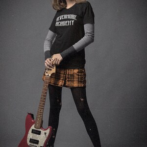 May include: A woman wearing a black t-shirt with the text "Nevermore Academy" and a plaid skirt is holding a red electric guitar.
