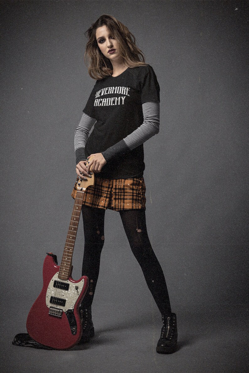 May include: A woman wearing a black t-shirt with the text "Nevermore Academy" and a plaid skirt is holding a red electric guitar.