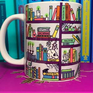 May include: White ceramic mug with a colorful illustration of a bookshelf filled with books. The books have titles like "Spells", "Love Potions", "Practical Magic", "Hamlet", and "How to Turn Men into Toads".