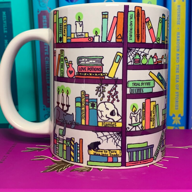 May include: White ceramic mug with a colorful illustration of a bookshelf filled with books. The books have titles like "Spells", "Love Potions", "Practical Magic", "Hamlet", and "How to Turn Men into Toads".