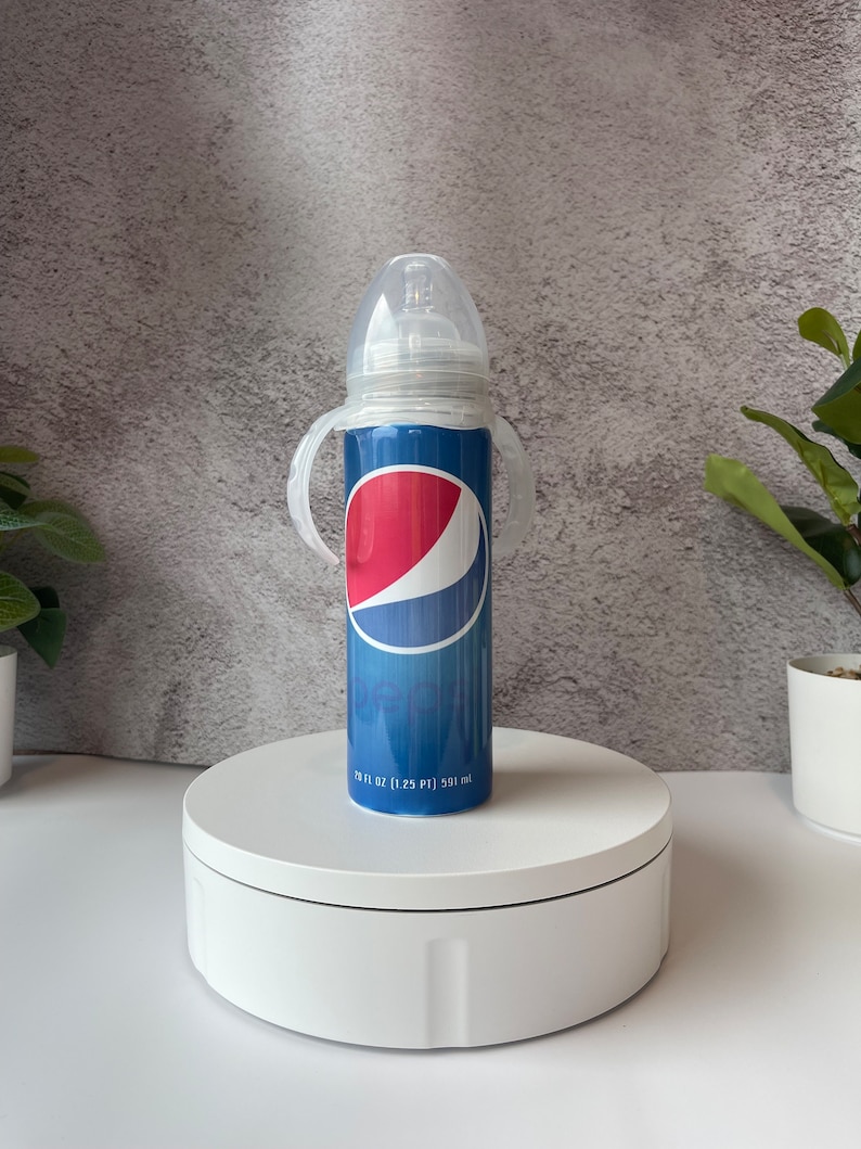 May include: A clear baby bottle with a blue and red Pepsi logo printed on the side. The bottle has two clear plastic handles and a clear silicone nipple. The bottle is sitting on a white turntable. The bottle is 1.25 pints or 591 ml.