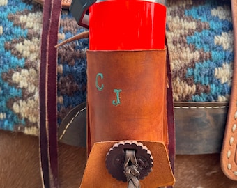 Op de afbeelding: Een bruine leren zadeltas met een rode en zilveren geïsoleerde waterfles erin. De tas heeft een bruine leren riem en een metalen gesp. De initialen "CJ" zijn gegraveerd op de voorkant van de tas.
