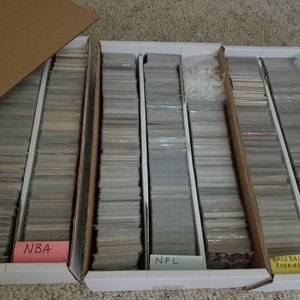 May include: Collection of sports trading cards organized in cardboard storage boxes. The boxes are labeled with pink, white, and yellow tags indicating the sports: NBA, NFL, and Baseball Rookies. The cards are stacked neatly.