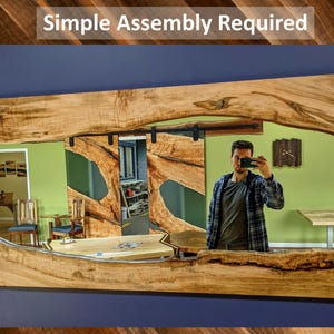 May include: A large wooden mirror with a live edge frame and a mirrored surface. The mirror is reflecting a room with a green wall, a wooden table, and a person taking a selfie.