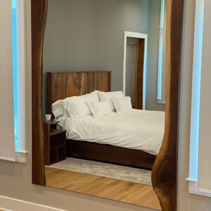 May include: A large, rectangular mirror with a natural wood frame. The frame has a unique, organic shape with a live edge. The mirror is reflecting a bedroom with a bed, nightstand, and a door.
