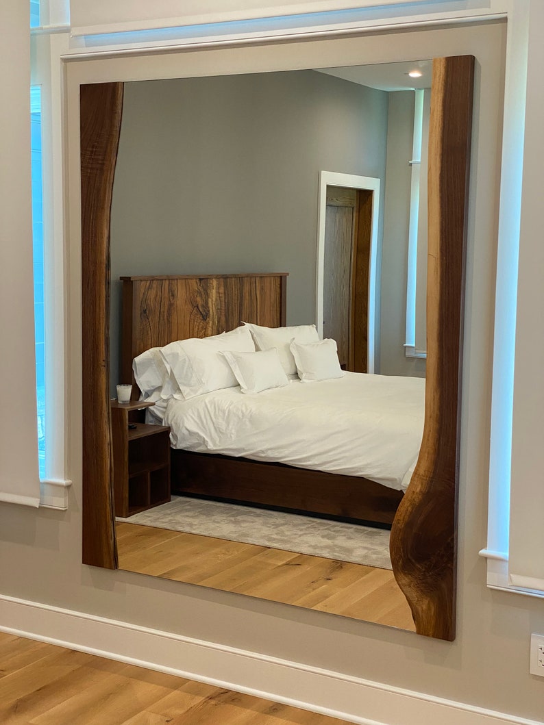 May include: A large, rectangular mirror with a natural wood frame. The frame has a unique, organic shape with a live edge. The mirror is reflecting a bedroom with a bed, nightstand, and a door.
