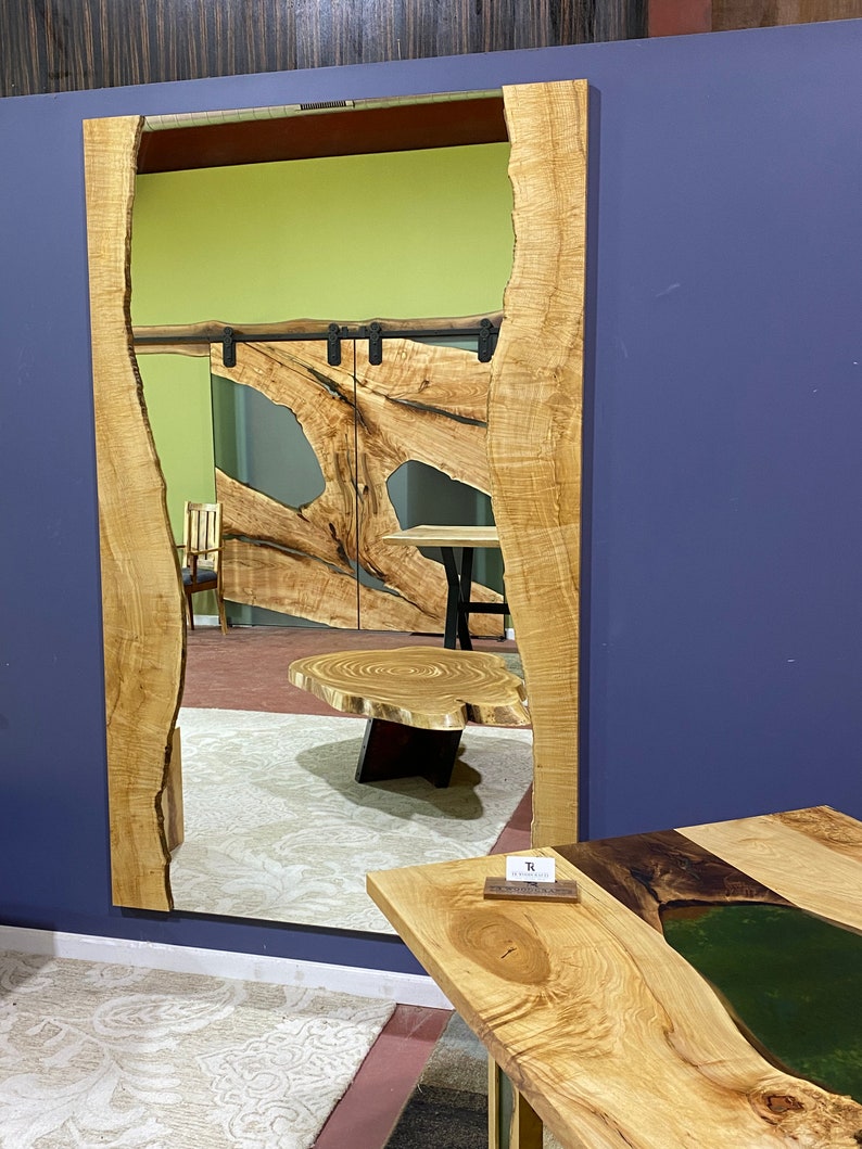 May include: A large, rectangular mirror with a natural wood frame. The frame has a unique, organic shape with a live edge. The mirror is reflecting a room with a wooden table and a rug.