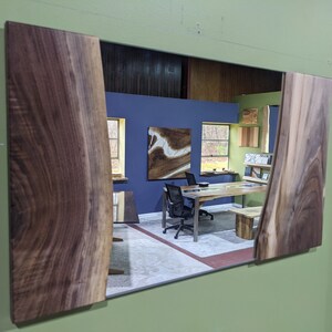 May include: A mirror with a wooden frame made of walnut wood with a live edge. The mirror reflects a room with a blue wall, a wooden table, and a chair.