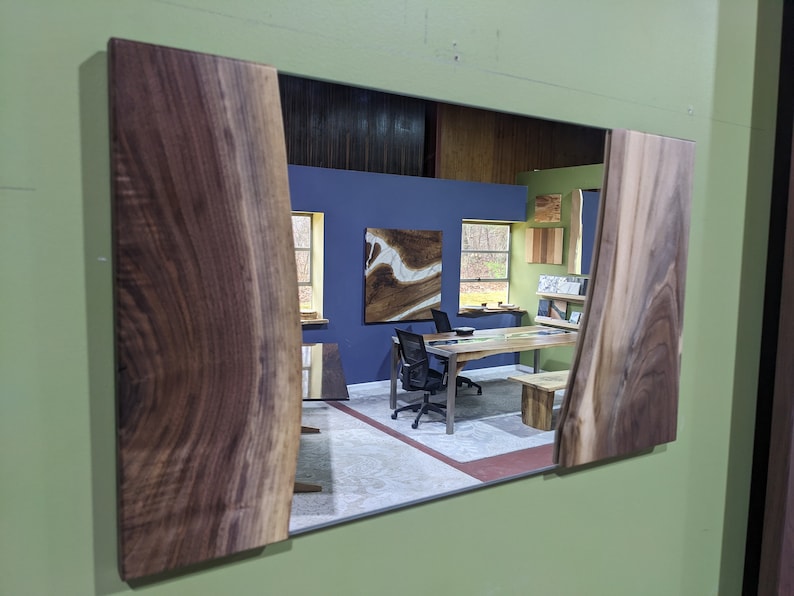 May include: A mirror with a wooden frame made of walnut wood with a live edge. The mirror reflects a room with a blue wall, a wooden table, and a chair.