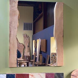 May include: A large rectangular mirror with a natural wood frame. The frame has a unique, irregular shape with a live edge design. The mirror reflects a room with wooden furniture and shelves.