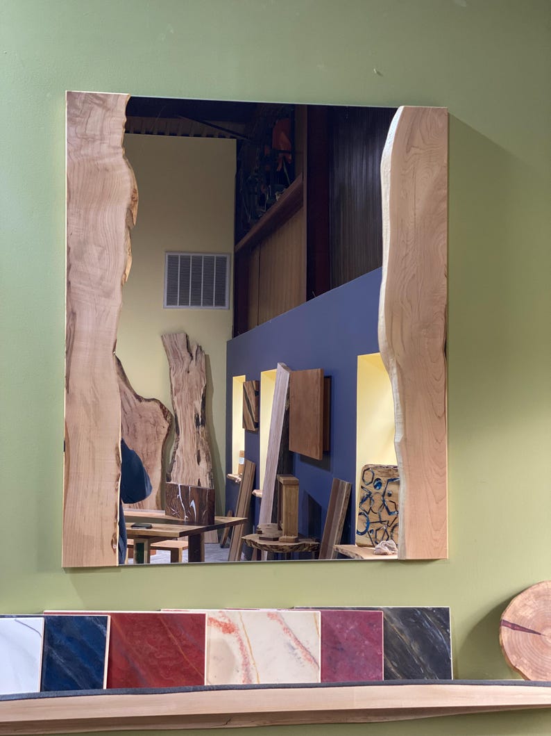 May include: A large rectangular mirror with a natural wood frame. The frame has a unique, irregular shape with a live edge design. The mirror reflects a room with wooden furniture and shelves.