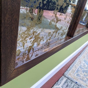 May include: A close-up of a dark brown wooden frame surrounding a mirror with a distressed gold finish. The mirror is mounted on a green wall with white trim.