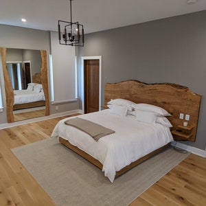 May include: A bedroom with a large wooden headboard, a white bed with white bedding, a brown blanket, and a small nightstand with a lamp. The room has hardwood floors and a large mirror on the wall.