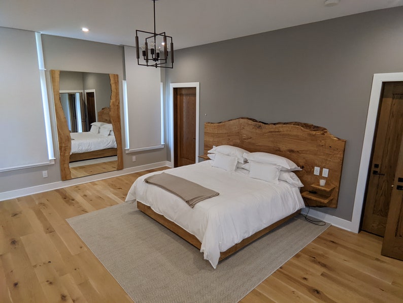 May include: A bedroom with a large wooden headboard, a white bed with white bedding, a brown blanket, and a small nightstand with a lamp. The room has hardwood floors and a large mirror on the wall.
