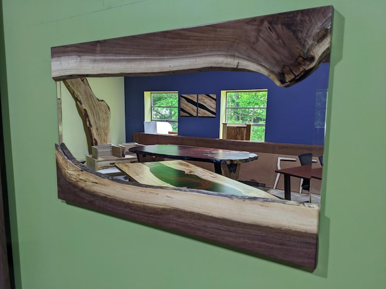 May include: A large wooden mirror with a live edge frame. The frame is made of walnut wood with a natural finish. The mirror reflects a room with a blue wall, two windows, and a table with a green epoxy river.