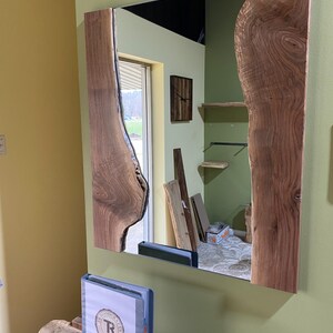 May include: A rectangular mirror with a wooden frame. The frame is made of two pieces of wood with a natural edge. The wood is a dark brown color. The mirror is reflecting a room with a wooden floor and a window.