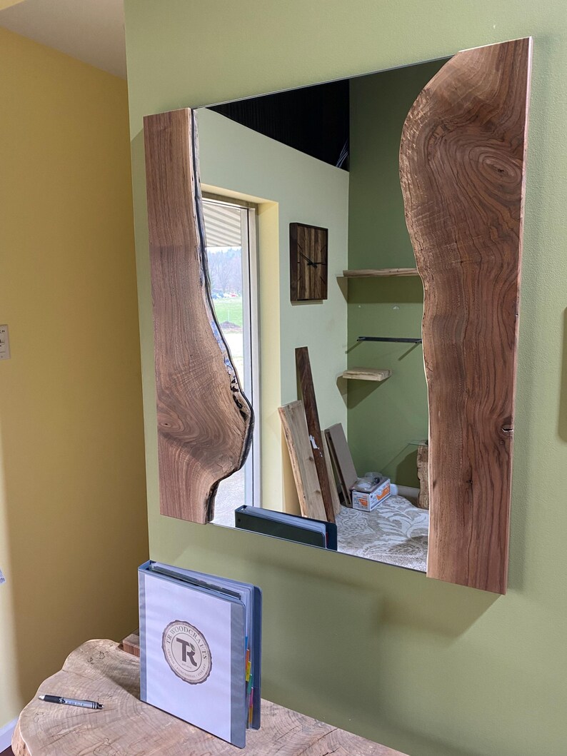 May include: A rectangular mirror with a wooden frame. The frame is made of two pieces of wood with a natural edge. The wood is a dark brown color. The mirror is reflecting a room with a wooden floor and a window.