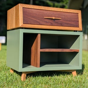 May include: A two-toned wooden nightstand with a drawer and open shelf. The drawer is made of dark brown wood with a light brown frame and a small wooden handle. The base is sage green with a walnut shelf and legs.