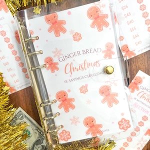 May include: A clear plastic binder with a gingerbread man Christmas theme. The binder has gold rings and a gold clasp. The cover has a white background with pink gingerbread men and snowflakes. The text on the cover reads "Gingerbread Christmas 1K Savings Challenge".